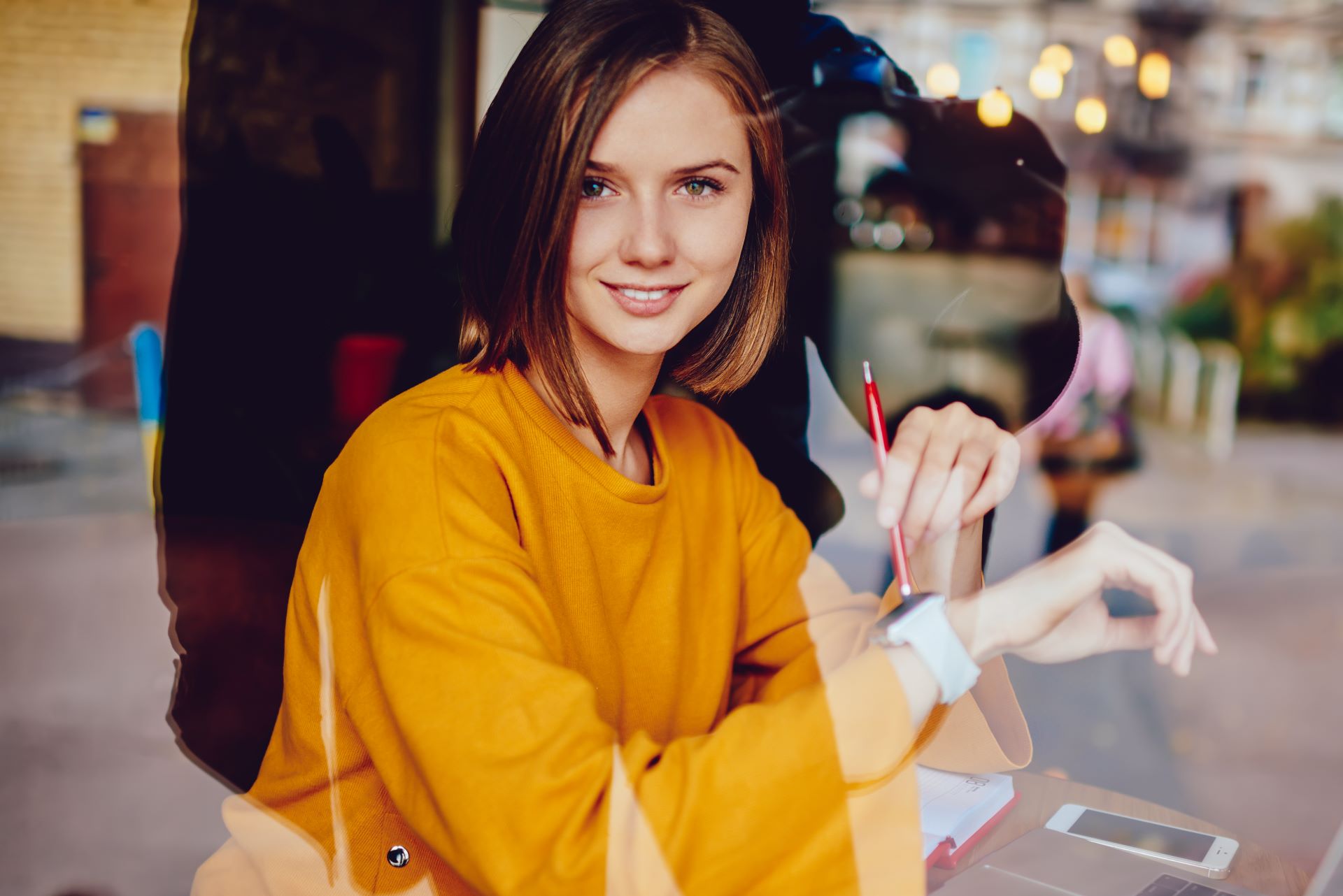 Eine junge Frau sitzt in einem modernen Büro oder Café und lächelt freundlich in die Kamera. Sie trägt einen gelben Pullover und hält einen Stift in der Hand, während sie durch eine Glasscheibe fotografiert wird. Im Hintergrund sind unscharf einige Lichter und urbane Details zu erkennen, die eine angenehme und entspannte Atmosphäre schaffen.