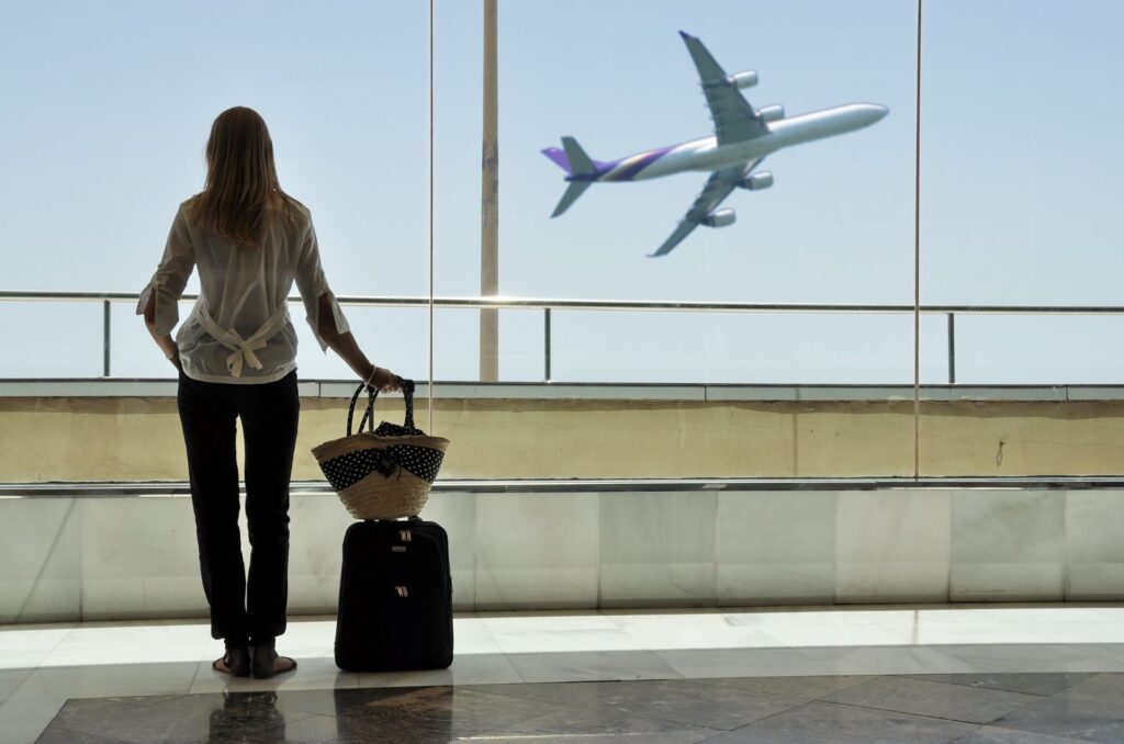 Reisende Frau mit Koffer blickt durch Fenster auf ein startendes Flugzeug