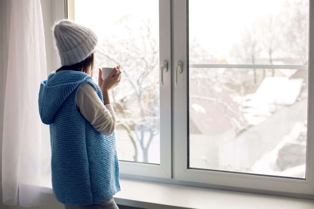 Frau mit warmer Kleidung blickt im Winter aus dem Fenster – Symbolbild für Heizbedarf und Energiepreise in NRW